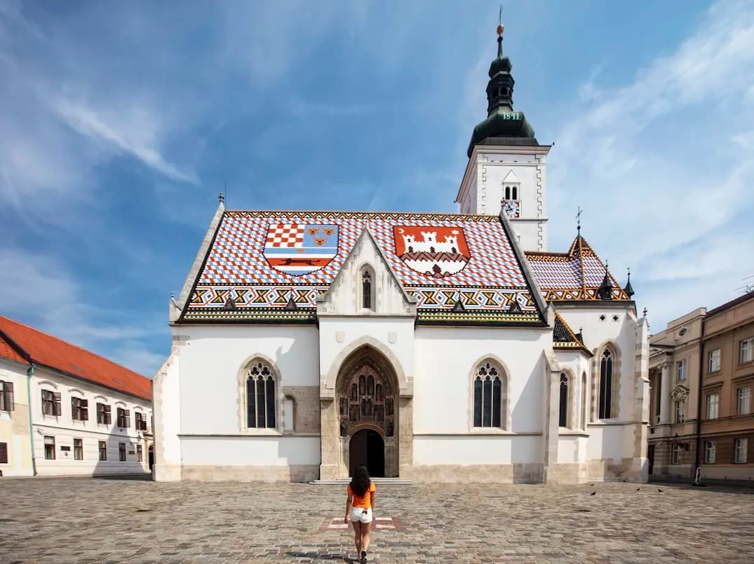 A lone traveler stands in front of St. Mark's Church, famous for its colorful tiled roof displaying the coats of arms of Croatia, Dalmatia, Slavonia, and Zagreb.