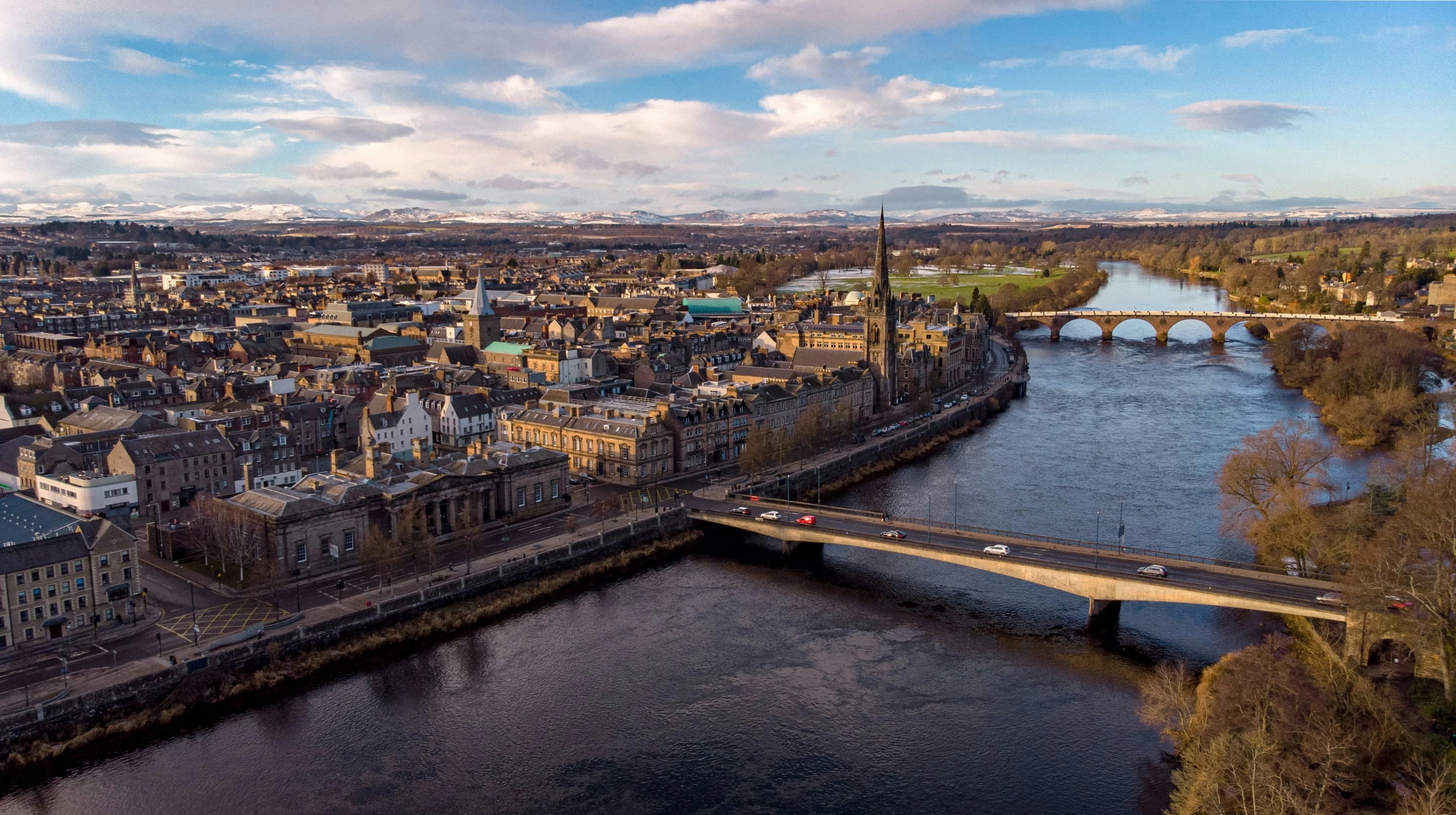 An aerial view of Perth and the River Tay, showing two bridges, the cityscape, and snow-capped mountains in the distance