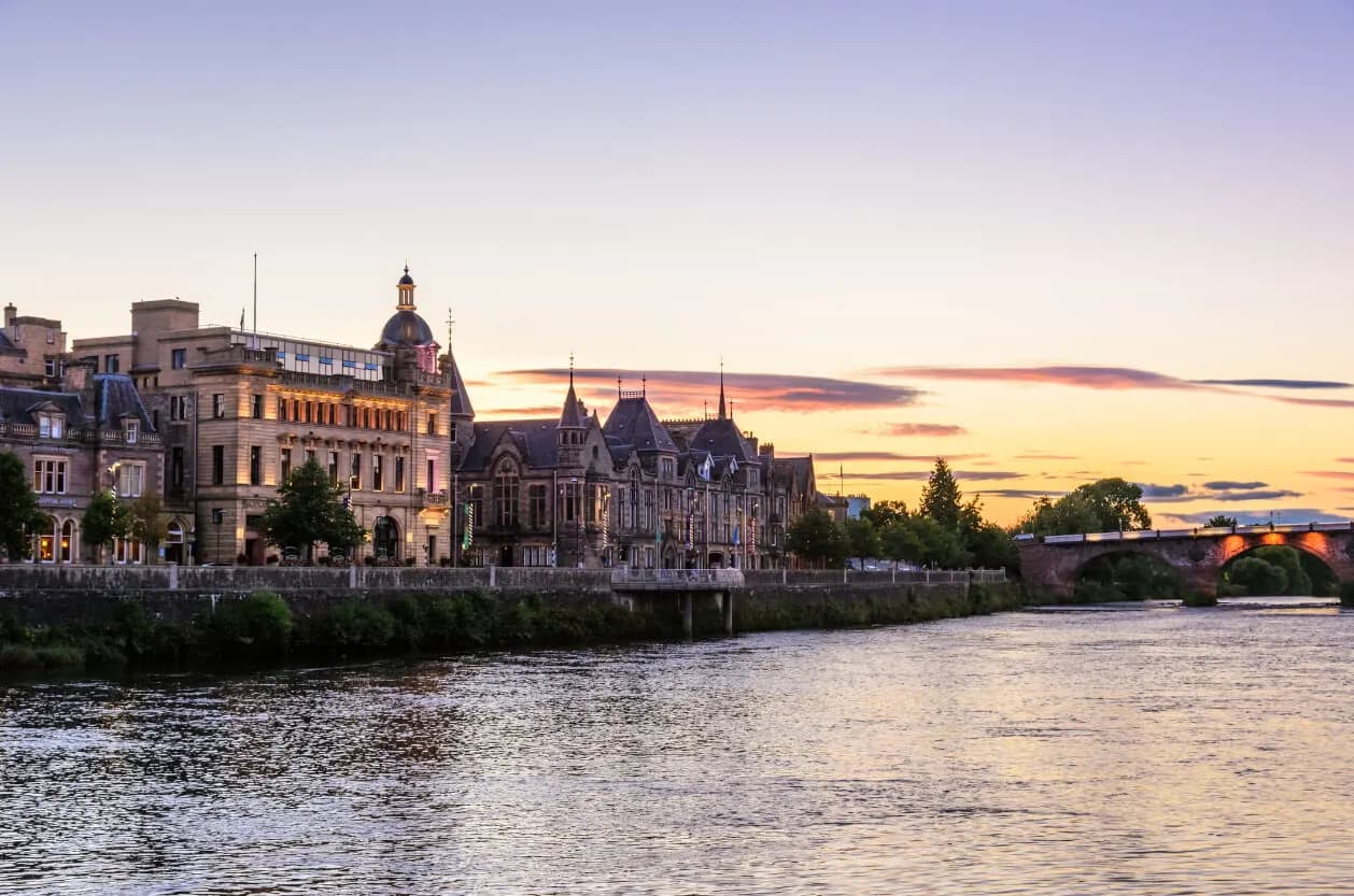 A sunset view of historic buildings lining the River Tay in Perth, with a stone arch bridge in the background