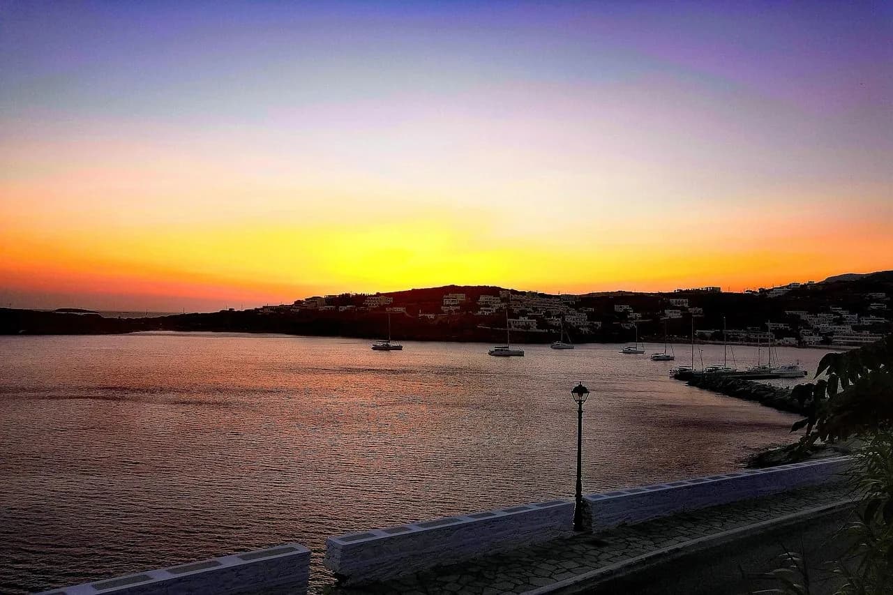 A scenic bay in Andros is bathed in the golden light of sunset, with boats anchored in the tranquil water and a town on the hillside.