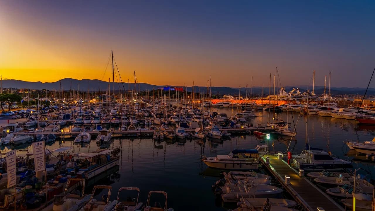 The port of Antibes is bustling with yachts and sailboats, with the city's lights and distant mountains in the background at sunset.