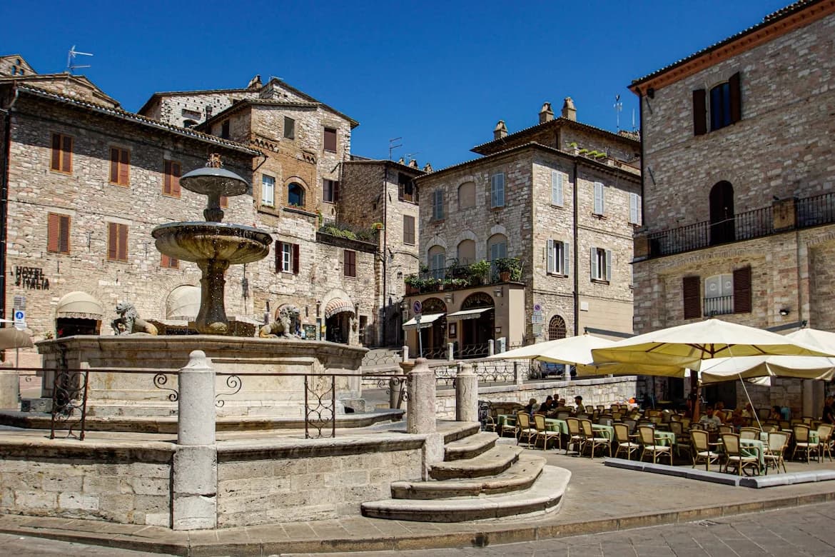 A large, historic fountain is the centerpiece of a cobblestone square in Assisi, with outdoor cafes and traditional stone buildings.