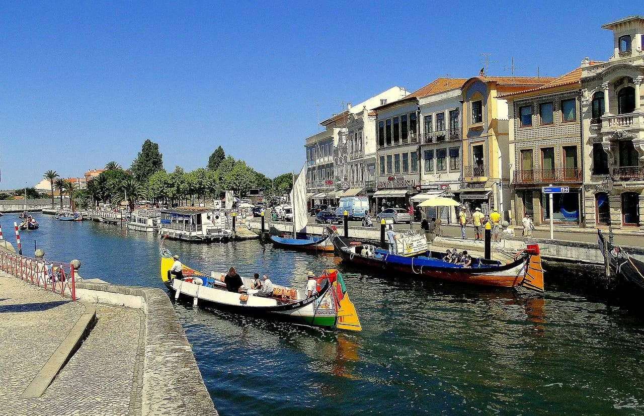 A Moliceiro boat, with its unique painted bow, is moored in a canal in Aveiro, with other boats and traditional buildings nearby.