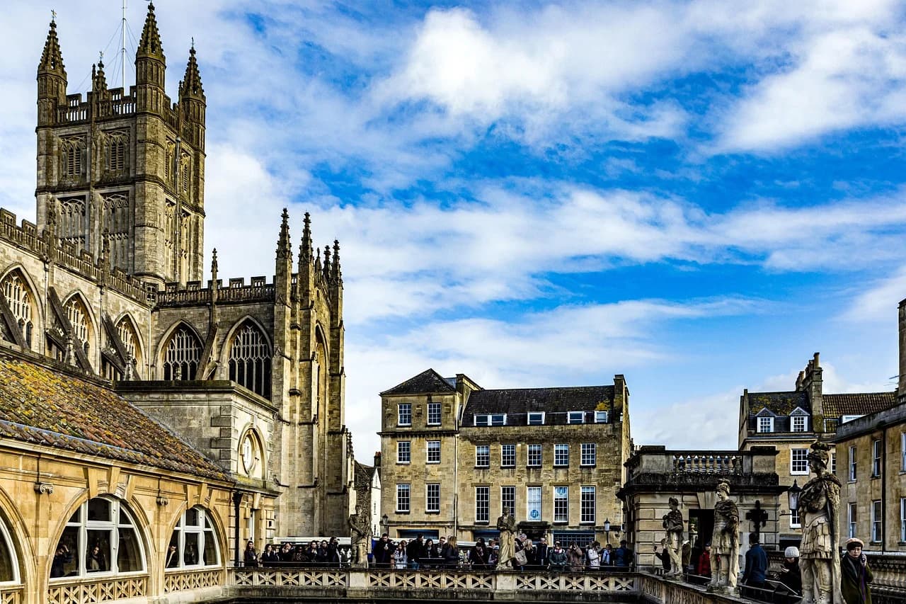 A crowd of people gathers around a historic fountain and statues outside the majestic Bath Abbey.