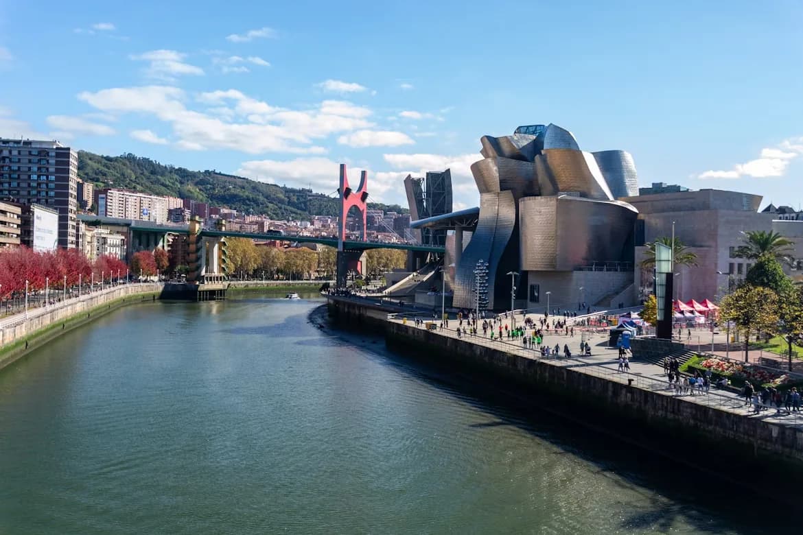 The iconic Guggenheim Museum Bilbao, with its futuristic, curved metal facade, stands next to a bridge over the Nervion River.