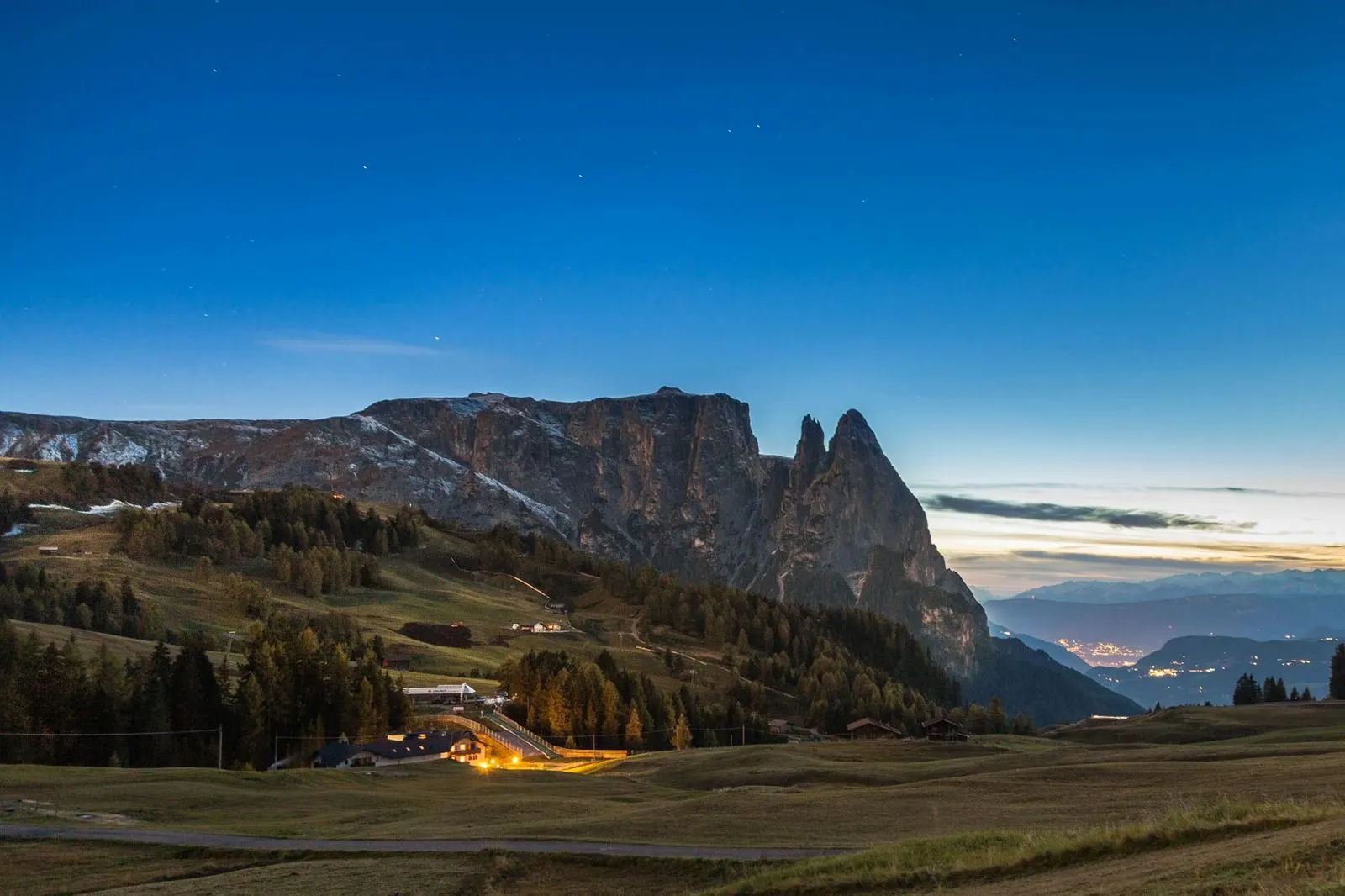 The dramatic, jagged peaks of the Dolomites stand tall against a twilight sky, with the lights of a distant village glowing below.