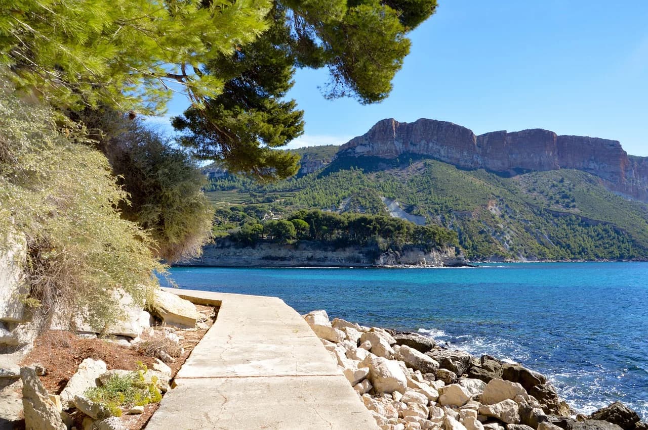 A peaceful stone pathway leads along the rocky coastline, with the blue sea and towering cliffs in the background.