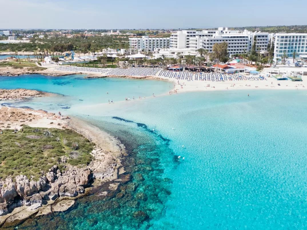An aerial view of a vibrant beach resort, with white buildings lining a long, white-sand beach and the sea in brilliant shades of blue.