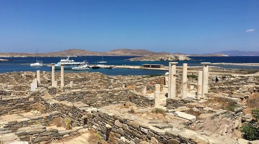 The ancient ruins of the Sanctuary of Apollo on Delos are set against the backdrop of a calm, blue harbor with moored boats.