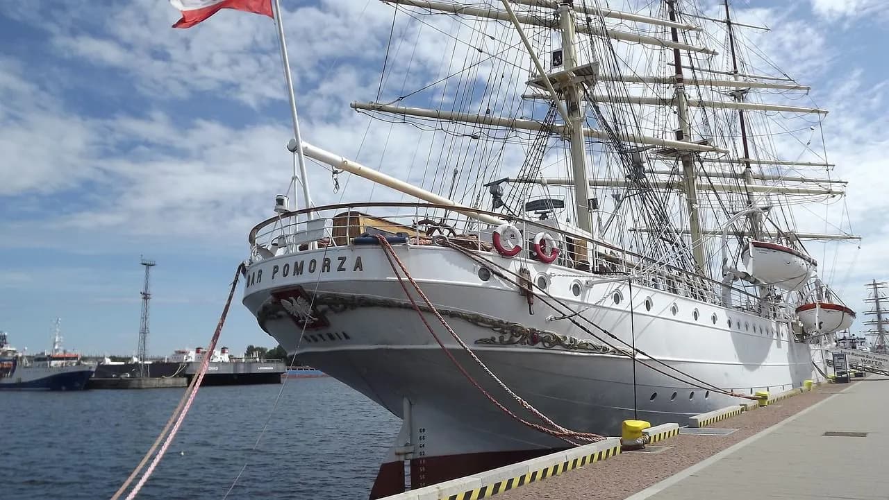 A tall, white sailing ship is docked at the harbor, with its rigging creating an intricate pattern against the blue sky.