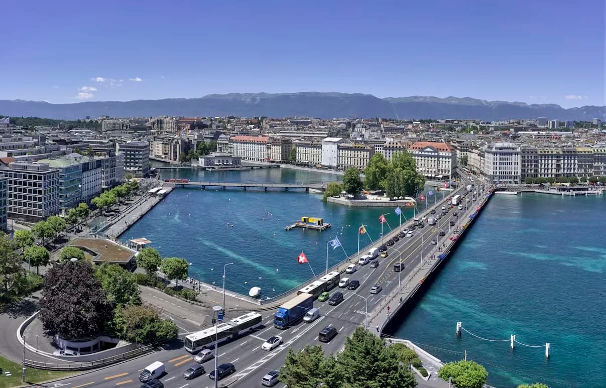 A high-angle view captures the city of Geneva, with the majestic Jet d'Eau fountain in the lake and the Swiss Alps in the distance.