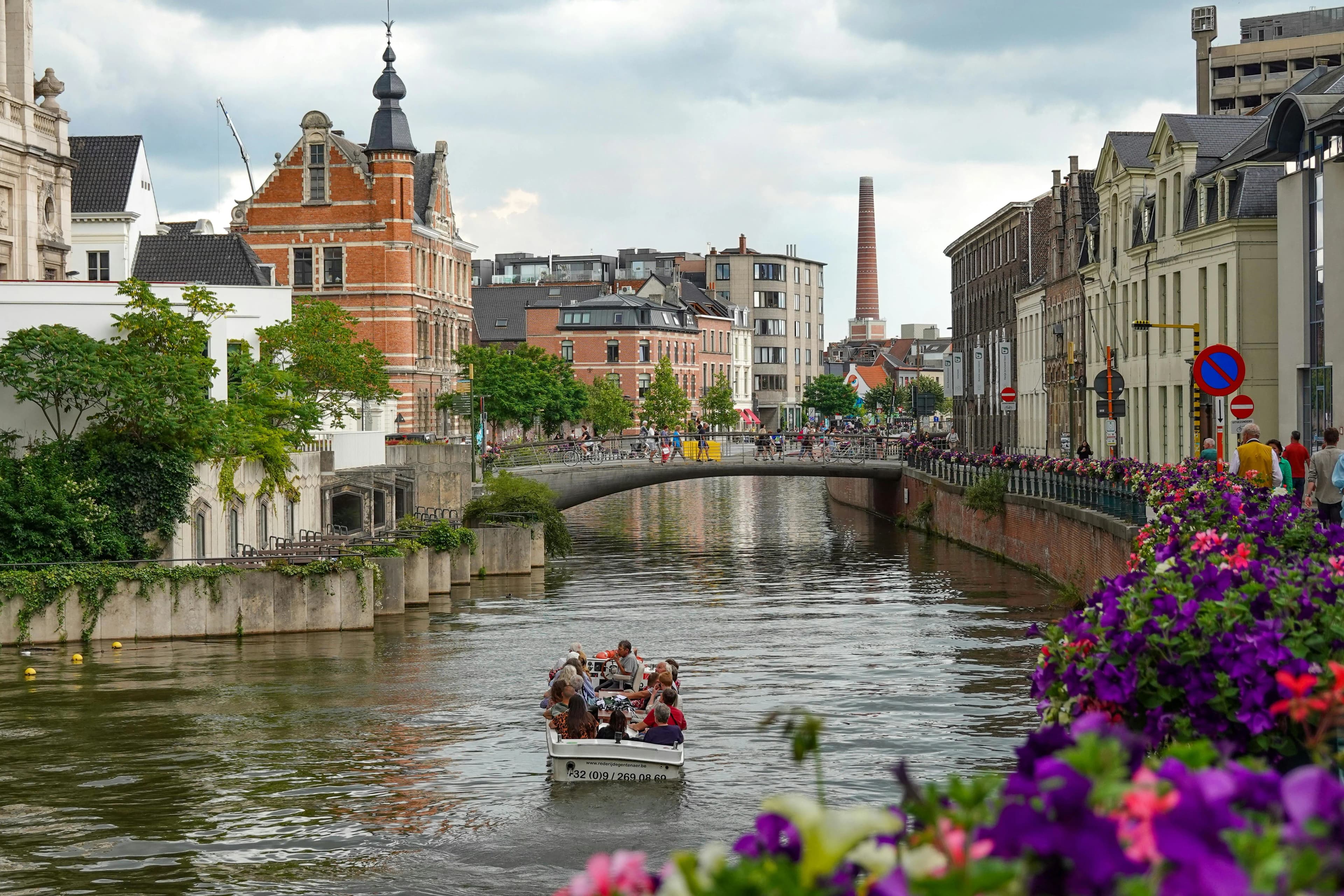 A tour boat filled with people cruises down a canal in Ghent, with beautiful flowers and historic buildings lining the waterway.