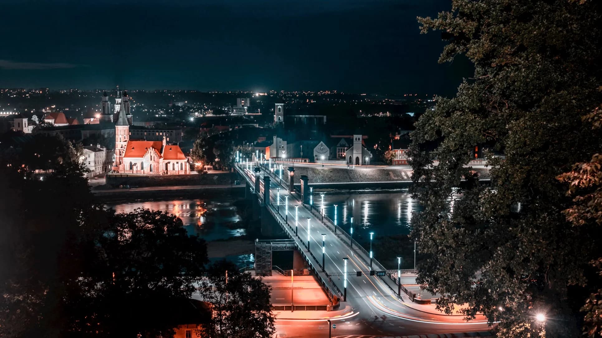 The city of Kaunas is illuminated at night, with the bright lights of a modern bridge reflecting on the river.