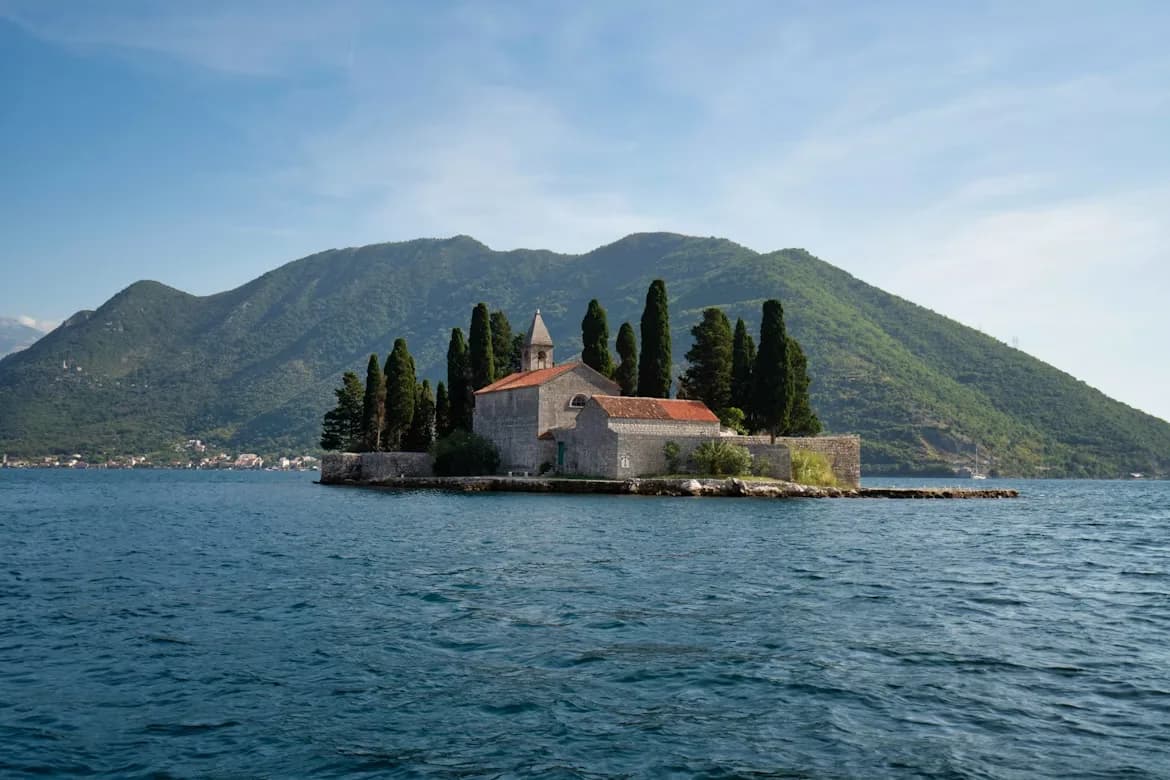 An iconic small island with a historic church sits in the calm waters of the Bay of Kotor, with the green mountains in the background.