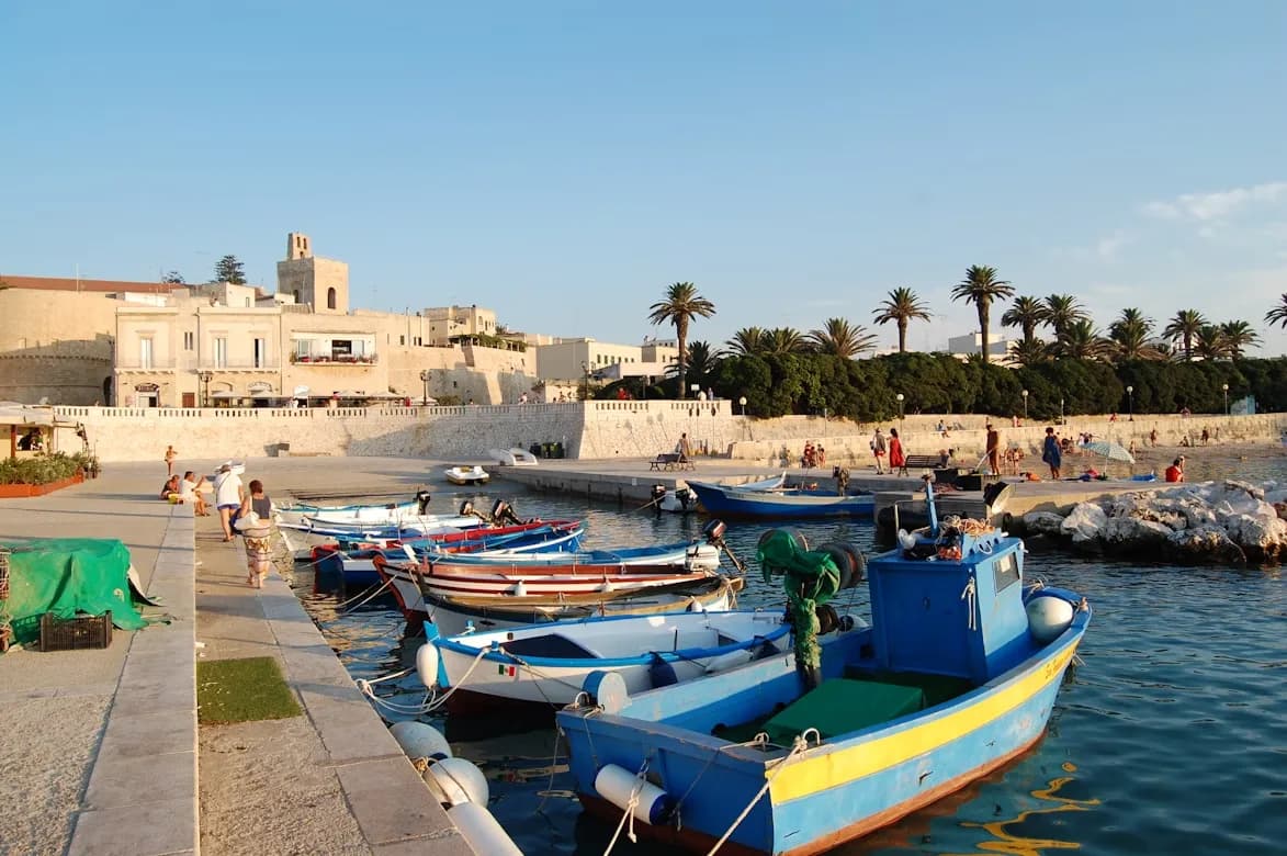 Fishing boats are moored in the tranquil harbor of Otranto, with a historic stone fortress and charming townscape in the background.