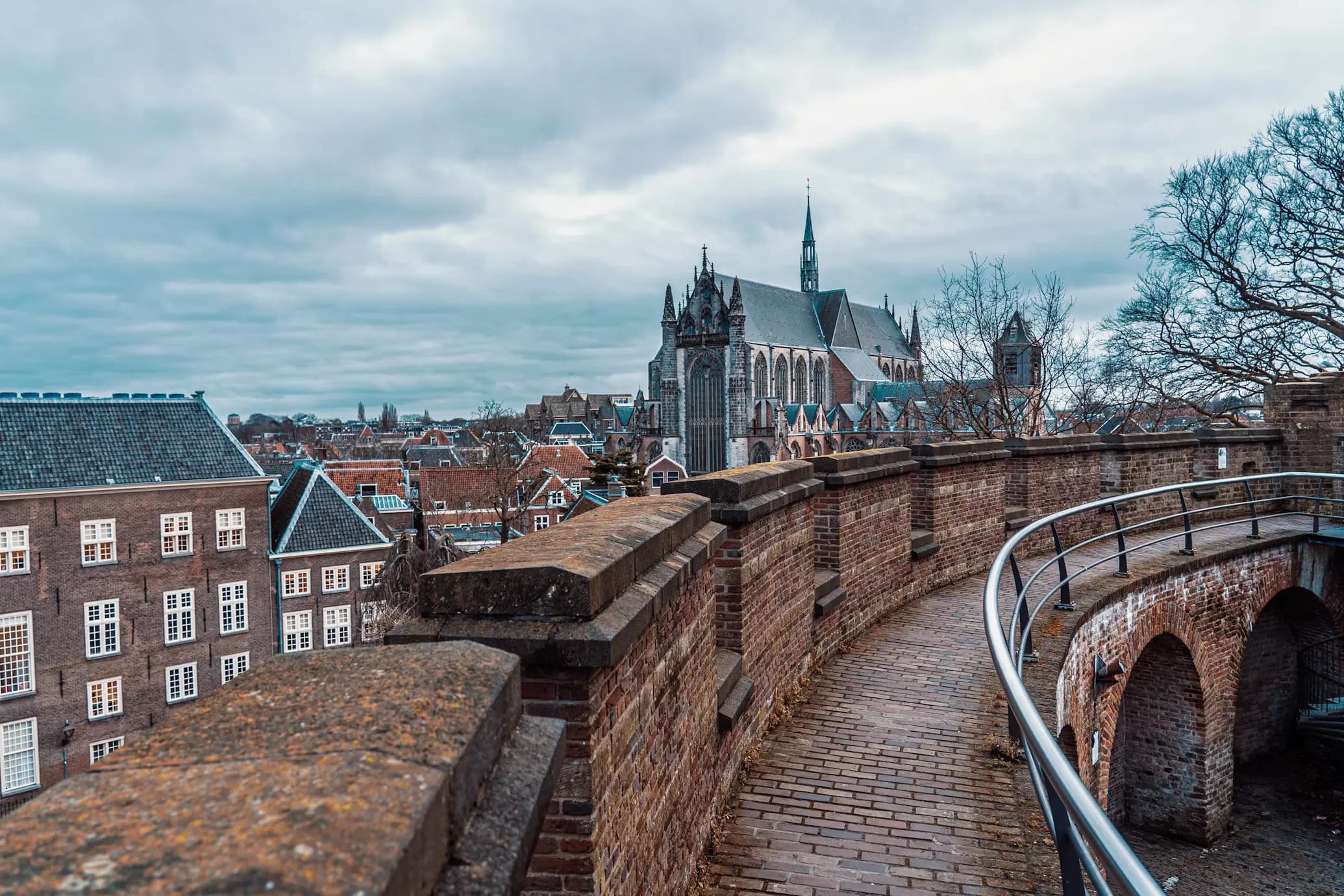 A medieval brick walkway on the ramparts of a fortress provides a view of the city, with a large Gothic church visible in the background.