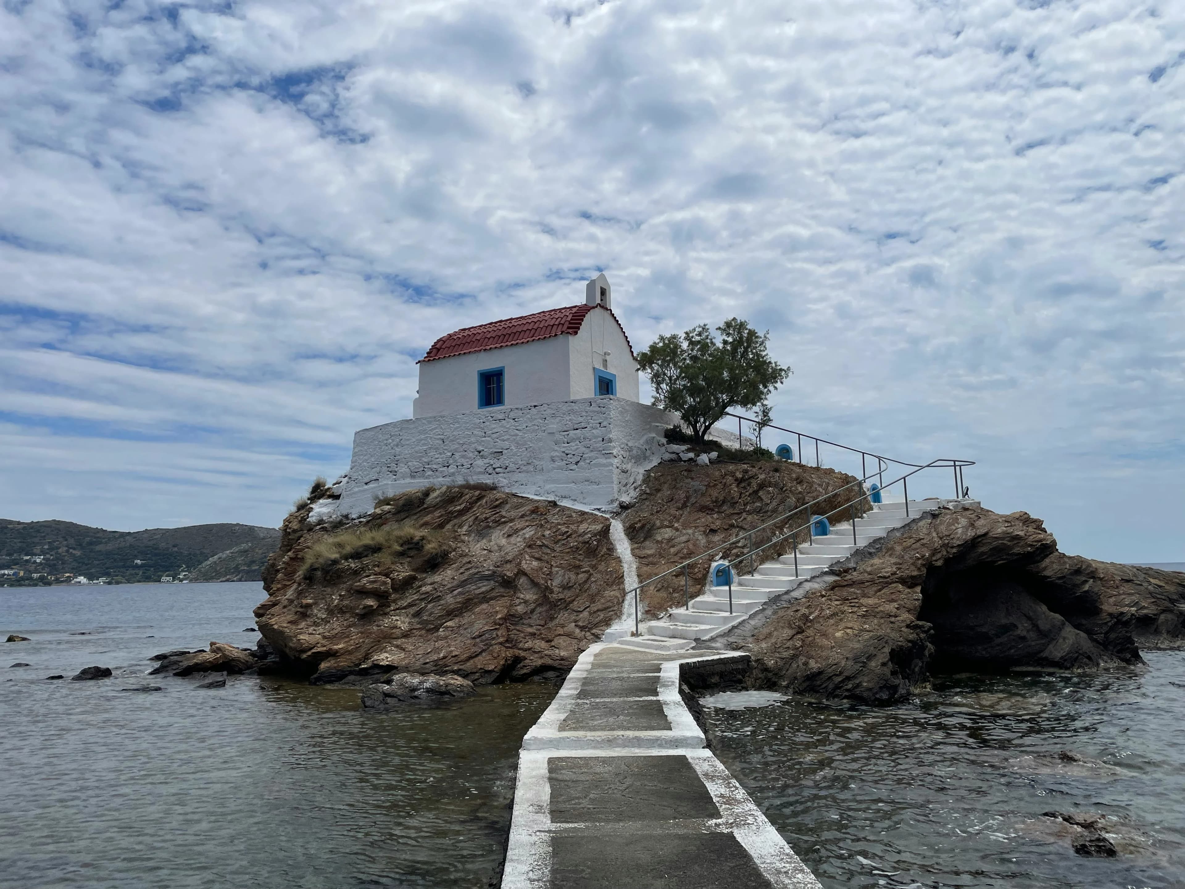 A small, picturesque white chapel with a red roof stands on a rocky islet, connected to the shore by a winding pathway and stairs.