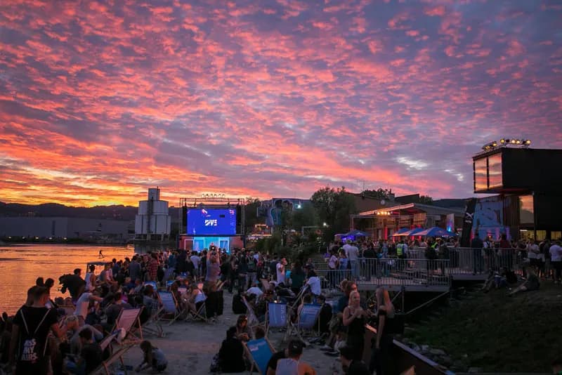 A crowd of people relaxes on a sandy beach by a river, watching a large screen under a dramatic sunset.