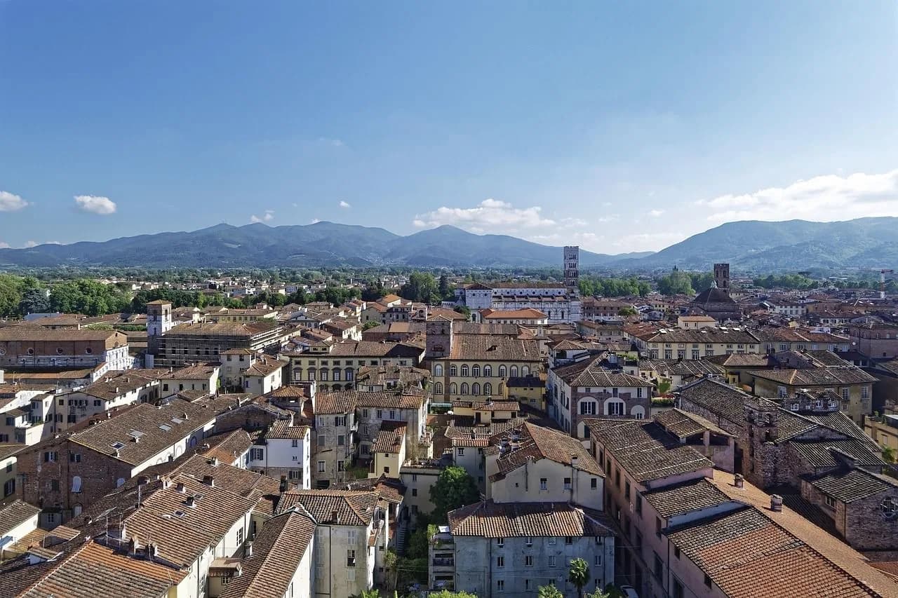 An elevated view captures the city's rooftops and historic bell towers, with lush green hills and mountains in the distance.