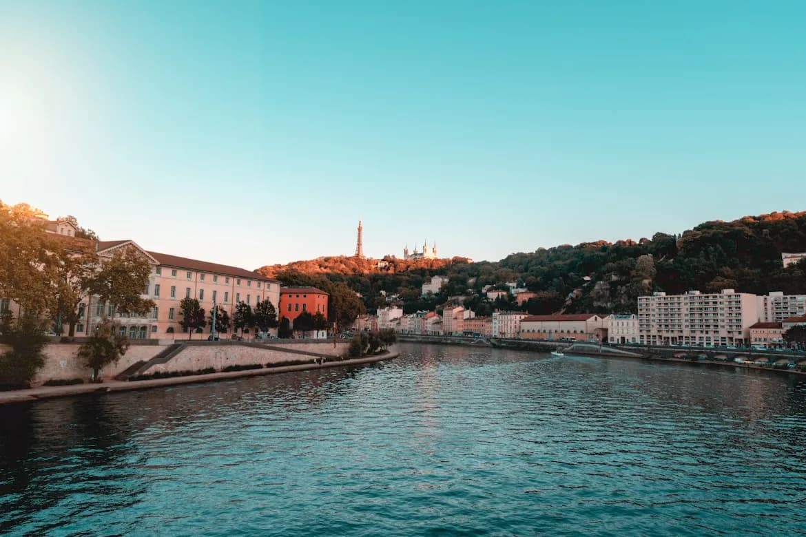 A serene river flows through the heart of Lyon, with historic buildings and the illuminated Basilica of Notre-Dame de Fourvière on the hill in the distance.