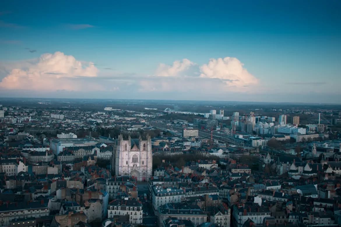 An aerial view captures the city of Nantes, with the gothic towers of the Nantes Cathedral and a wide river flowing in the background.