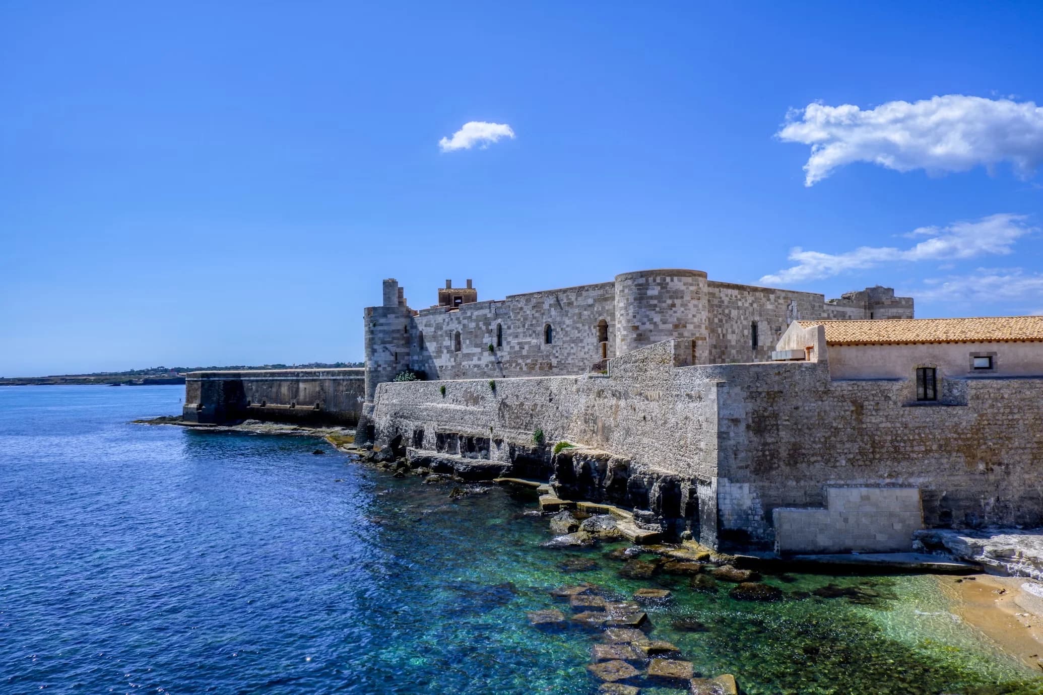 The historic Maniace Castle, a medieval fortress, stands on the edge of the sea, with its stone walls rising from the clear blue water.