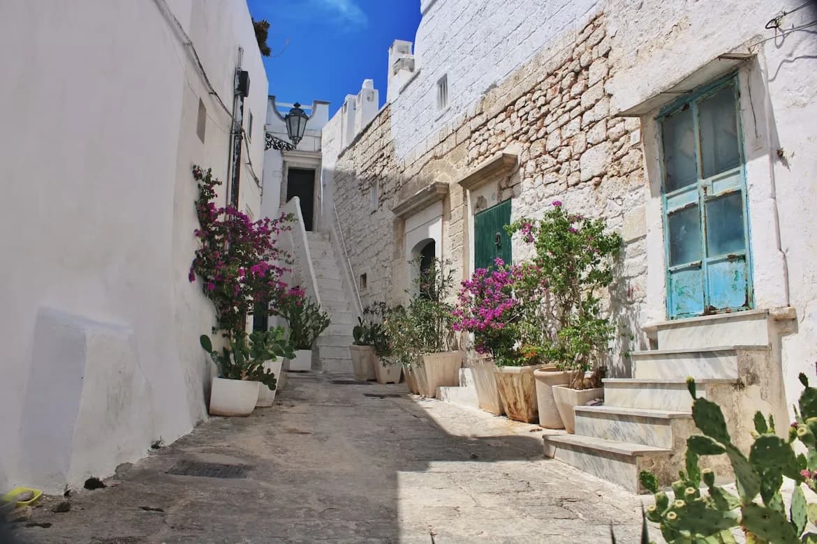 A narrow, cobblestone alleyway in Ostuni is lined with traditional white-washed buildings and vibrant flower pots.