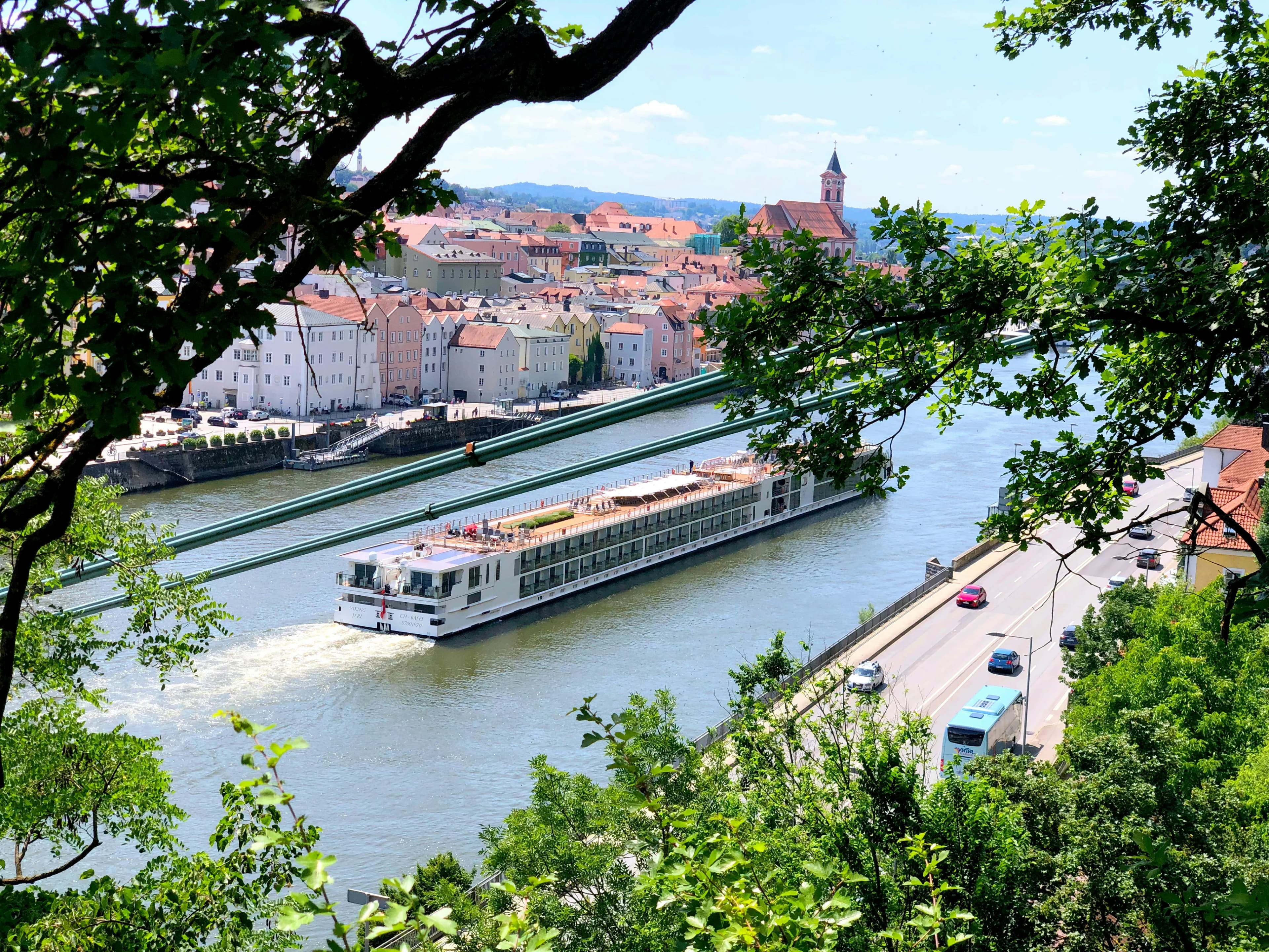 A long, narrow tour boat glides down the Danube River, with the historic city of Passau on a hill in the background.