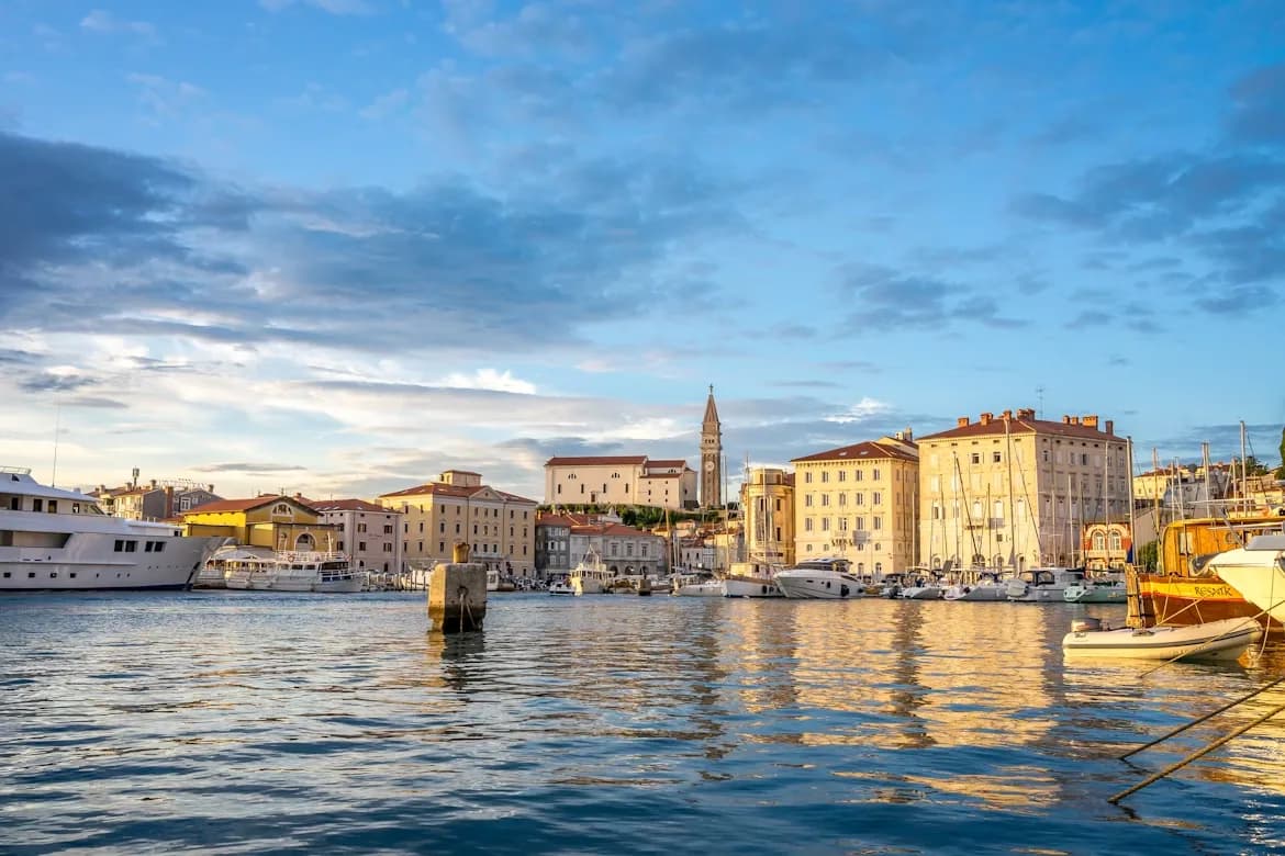Yachts and sailboats are moored in the harbor of Piran, with the historic town and the bell tower of St. George's Church in the background at sunset.