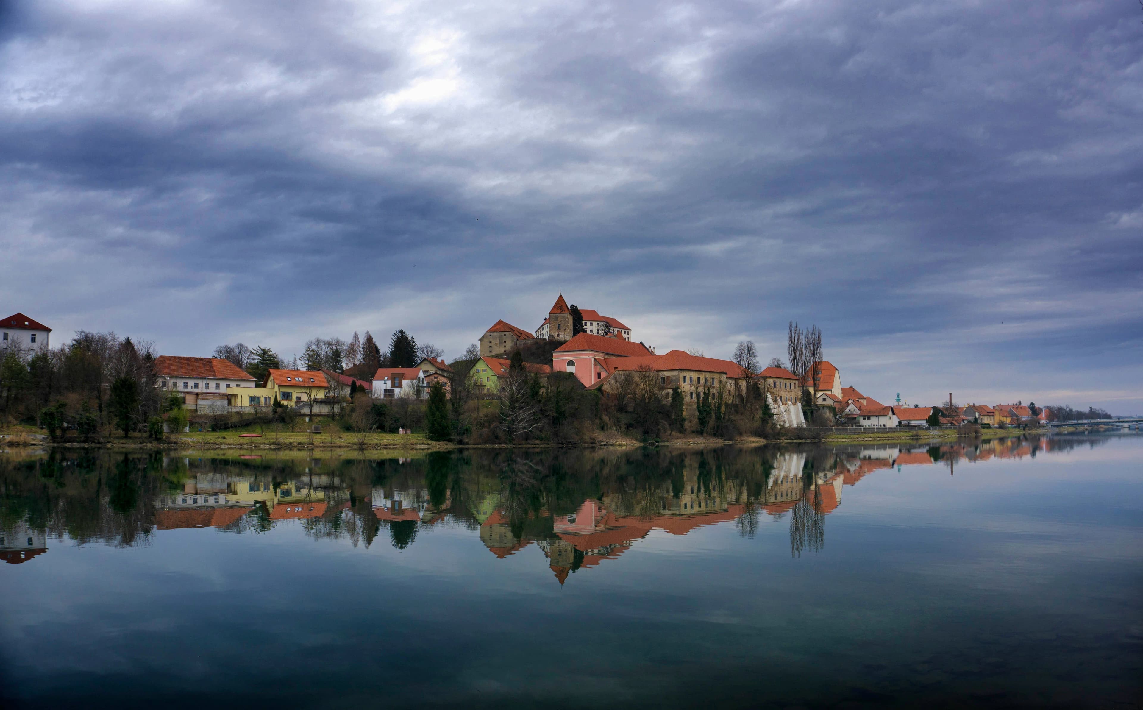 The historic Ptuj Castle, a medieval fortress, is beautifully reflected on the calm waters of the Drava River under a cloudy sky.