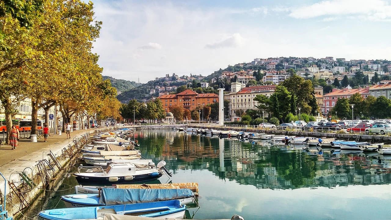A bustling harbor in Rijeka is filled with moored boats, with the city's historic buildings climbing the hillside in the background.