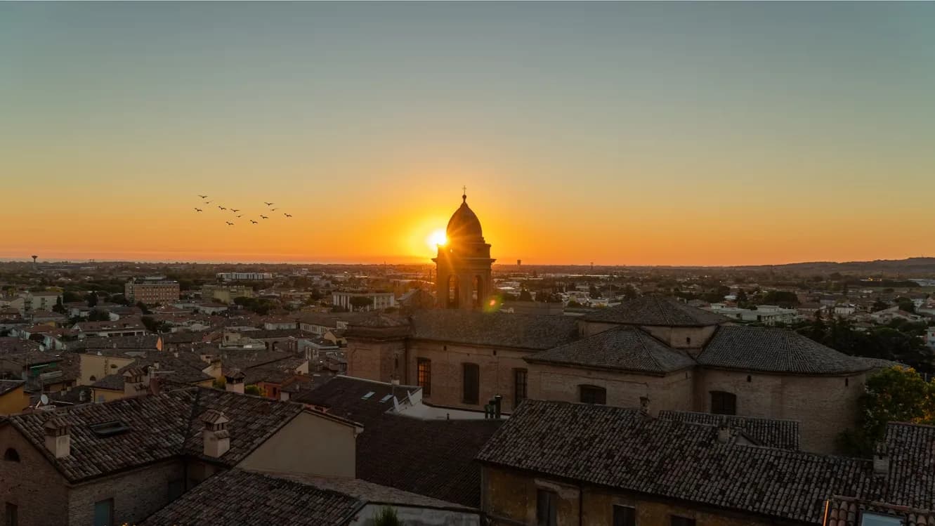 The sun sets behind a historic church bell tower, casting a golden glow over the rooftops of Rimini.
