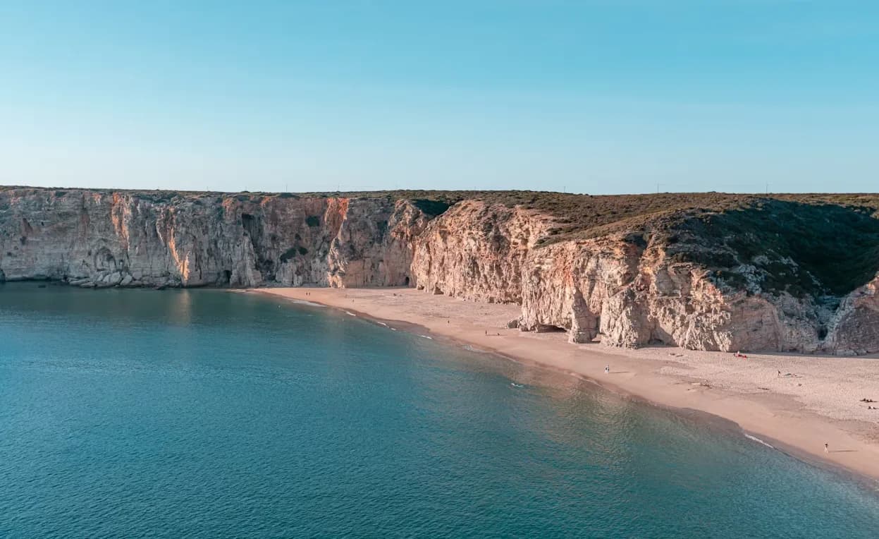 A secluded sandy beach is framed by dramatic, rocky cliffs, with the clear blue ocean stretching to the horizon.