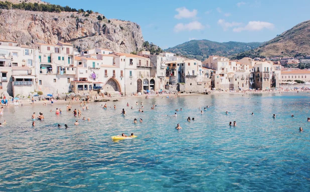 People swim and relax on a sandy beach, with the historic town of Cefalù and a towering cliff in the background.