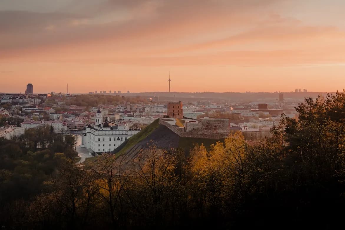 The historic Gediminas Castle Tower stands on a hill, overlooking the rooftops of Vilnius' Old Town at sunset.