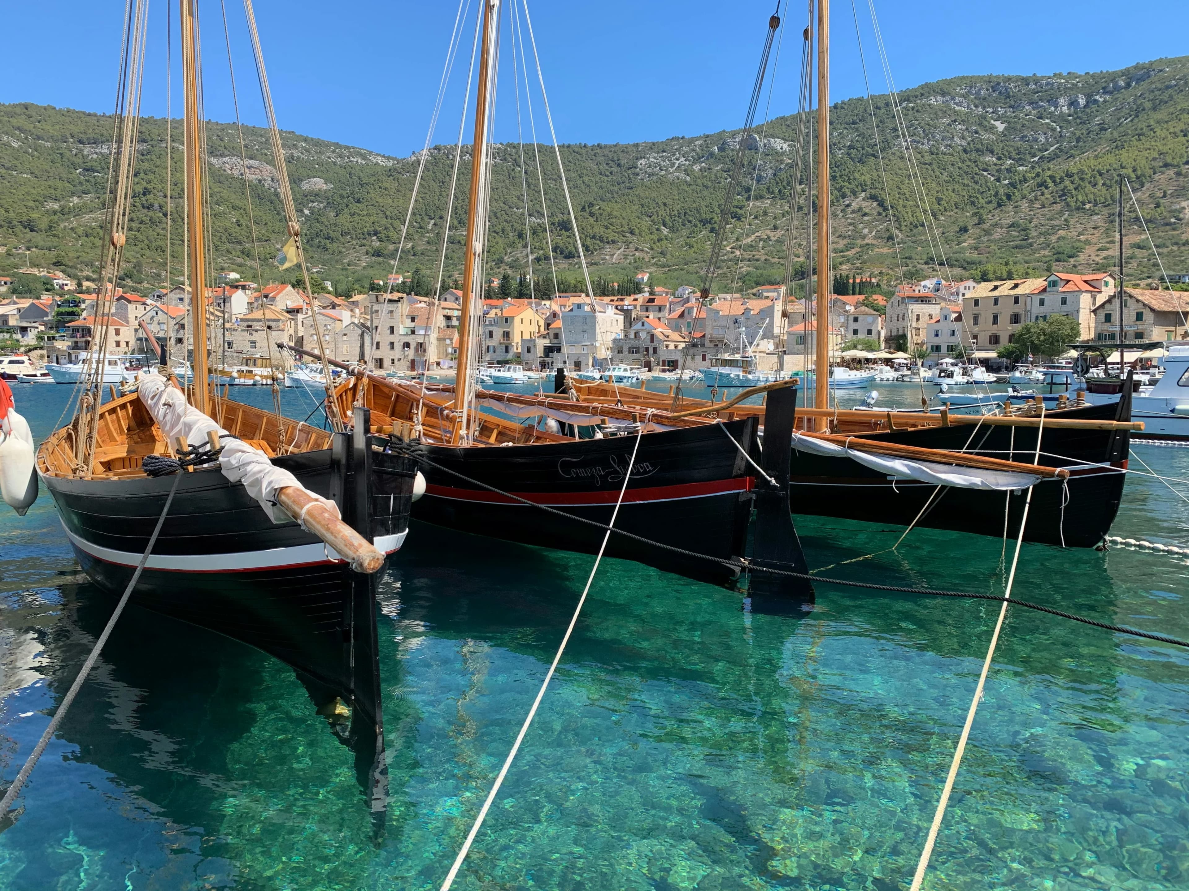 Three traditional wooden sailboats are moored in a bay with clear turquoise water, with the colorful buildings of a coastal town in the background.