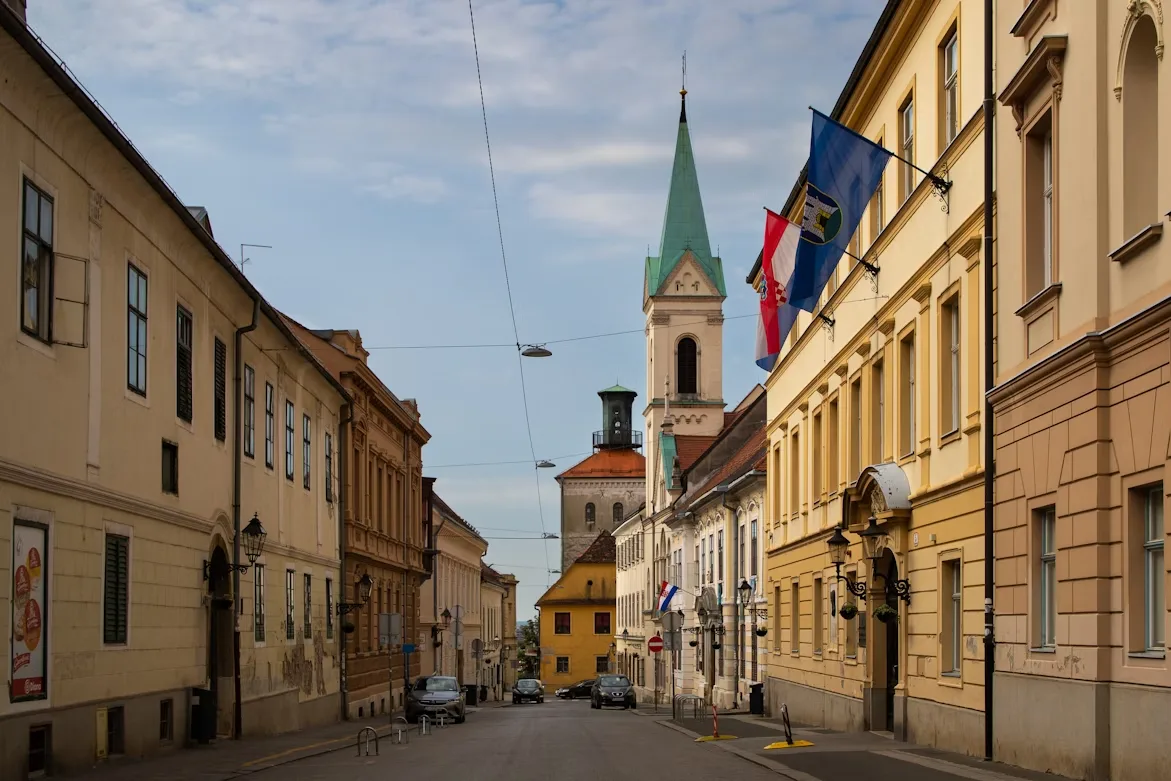 A historic, narrow street in Zagreb's Upper Town is framed by classic buildings, with a prominent church steeple visible in the distance.