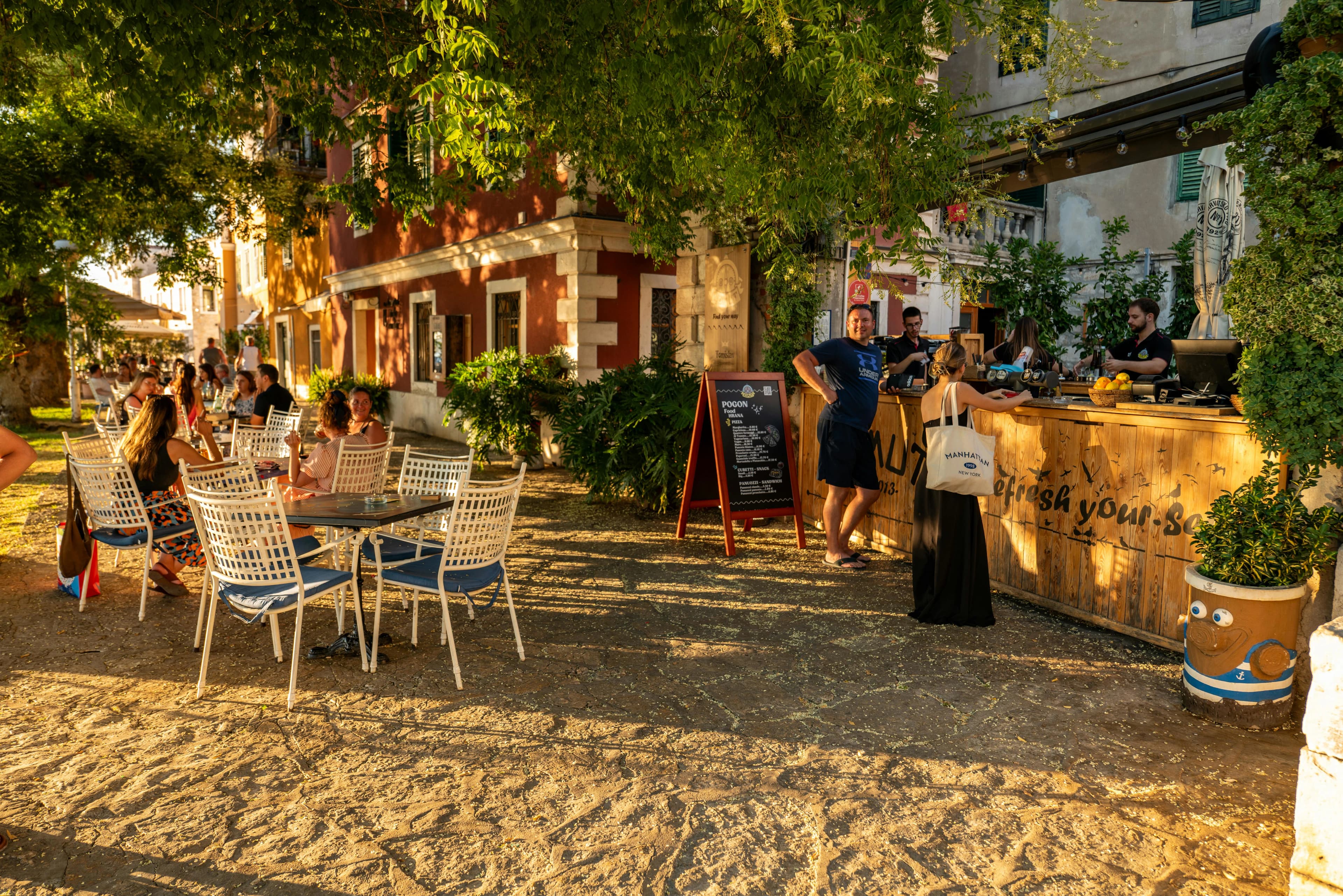 People dine at outdoor tables in a sun-drenched square, with a large, leafy tree providing shade and a historic building in the background.
