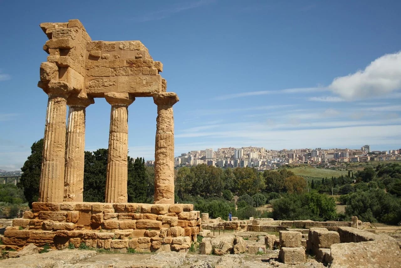 The ruins of a Greek temple with its original columns stand in the foreground, with the modern city of Agrigento on a hill behind it.