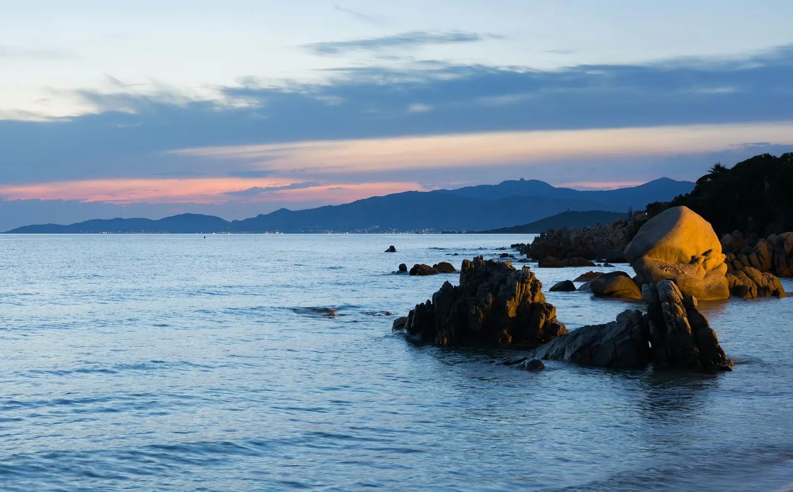 A rocky beach with large, smooth stones is bathed in the warm light of a sunset, with the sea stretching toward the mountains in the distance.