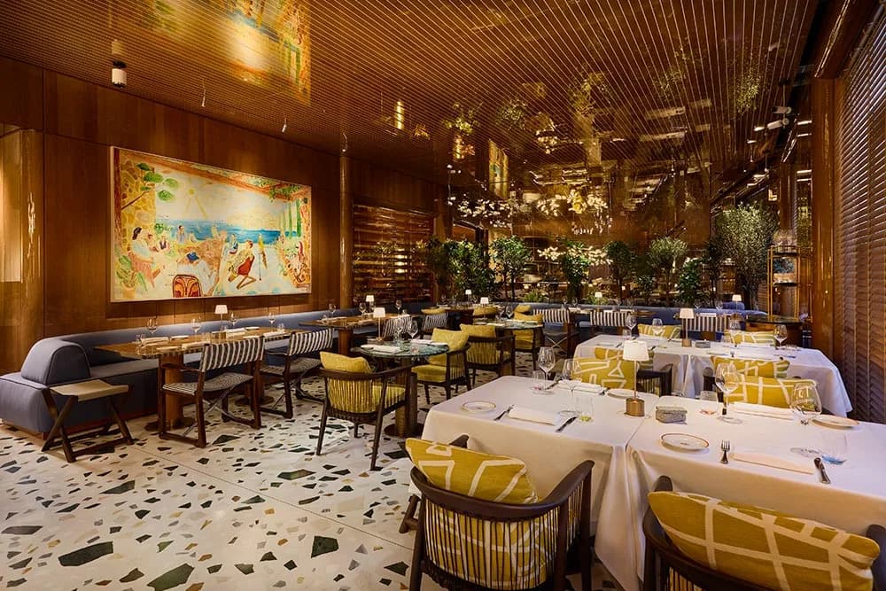 A modern restaurant with a large, colorful mural on the wall is filled with tables and chairs, with a unique chandelier made of small lights.