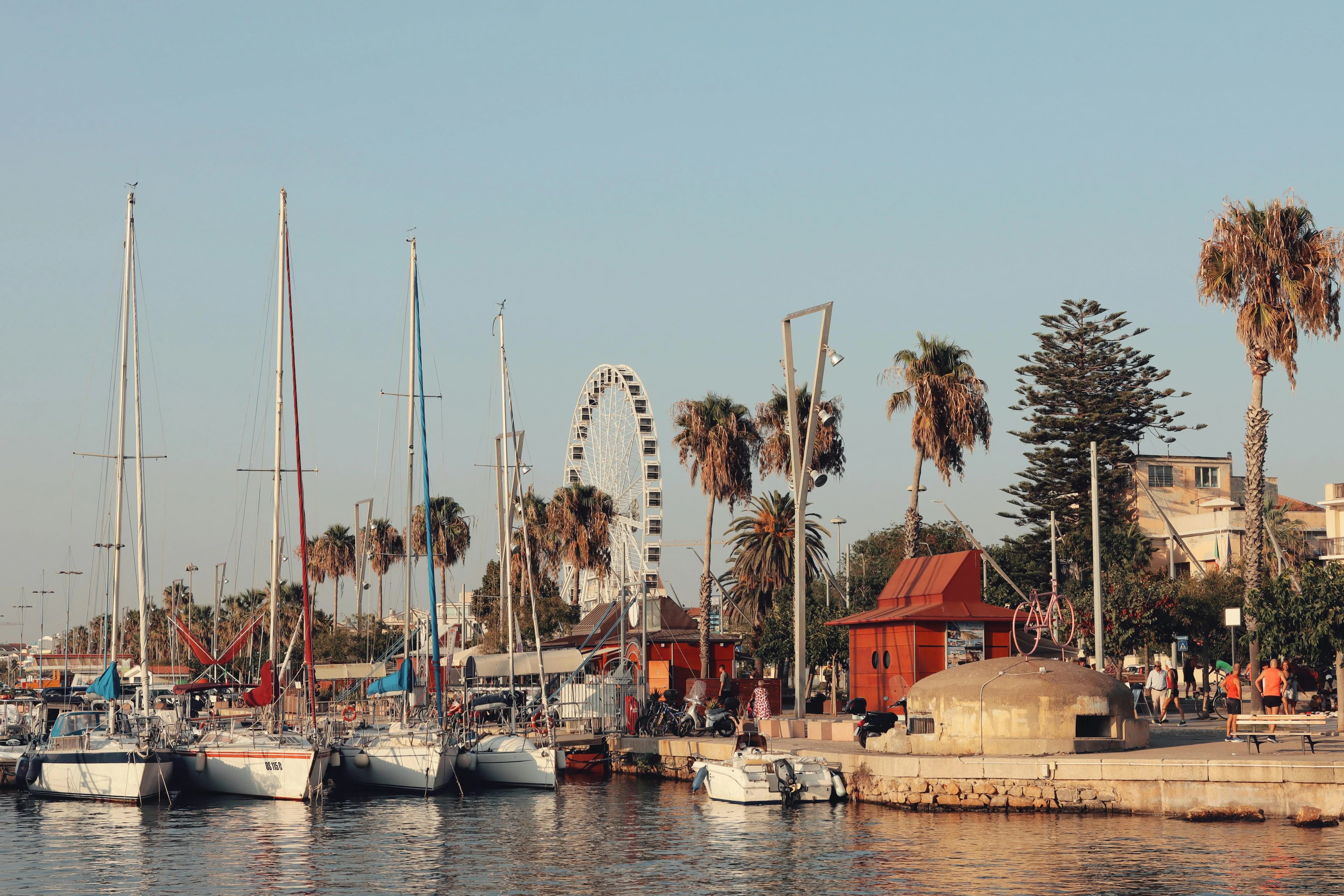 A large Ferris wheel and palm trees stand behind a row of sailboats and yachts moored in the harbor of Alghero.