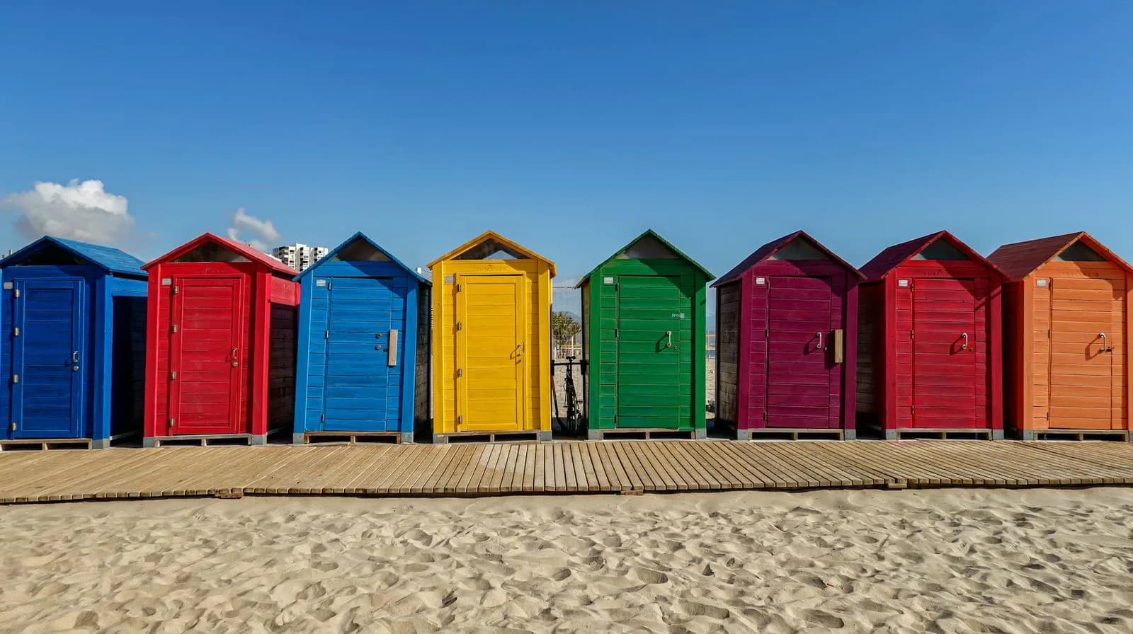 A row of colorful wooden beach huts, painted in a rainbow of hues, stands on a sandy beach.
