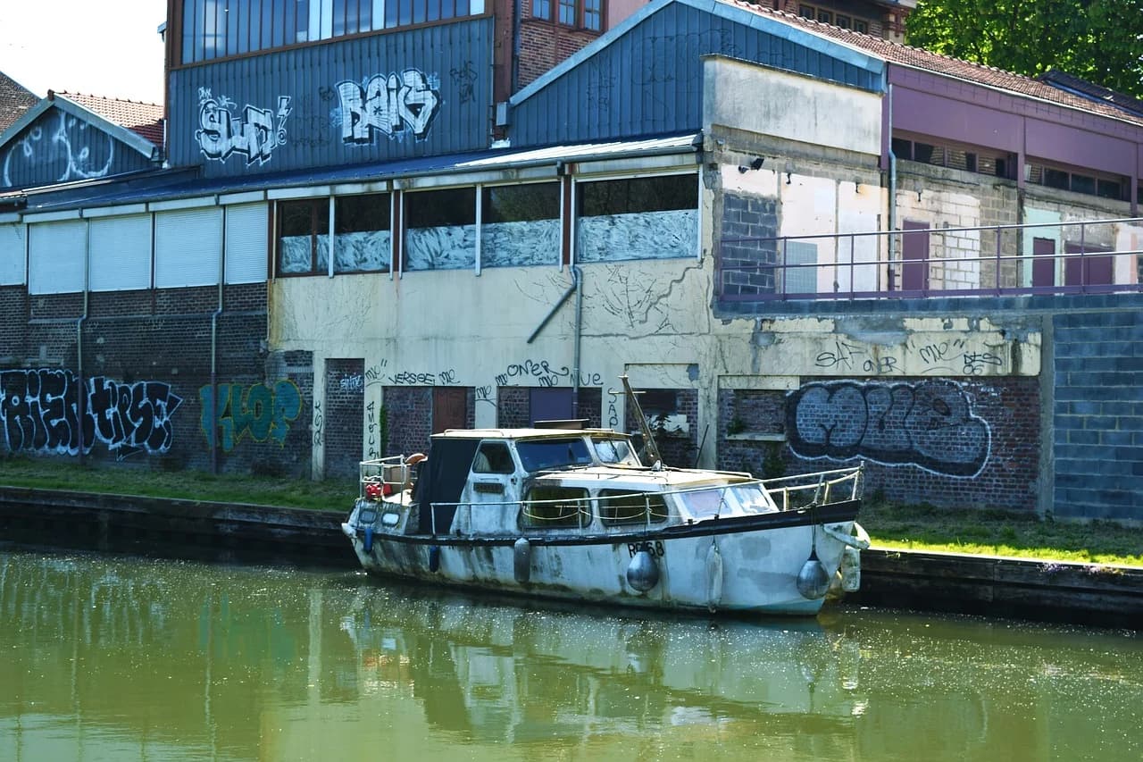 A weathered boat is moored in a canal, with graffiti-covered buildings on the opposite bank.