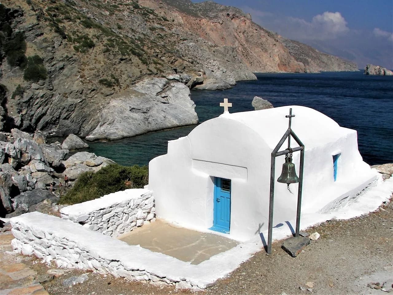 A small, white chapel with a bright blue door and a bell hangs on a stand in a rocky cove by the sea.