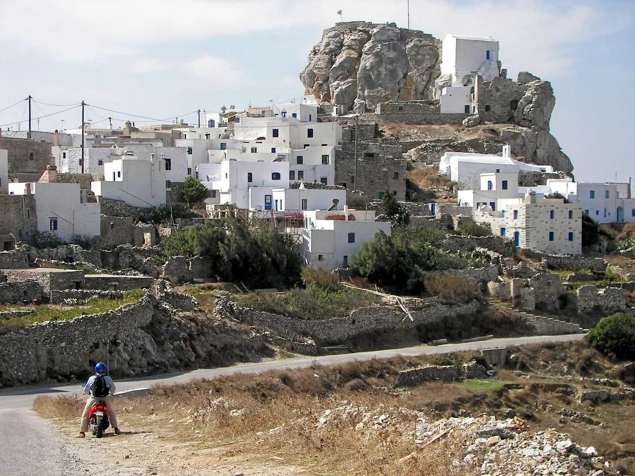 A person on a scooter is parked on a road overlooking a traditional white-washed town on a rocky hill.