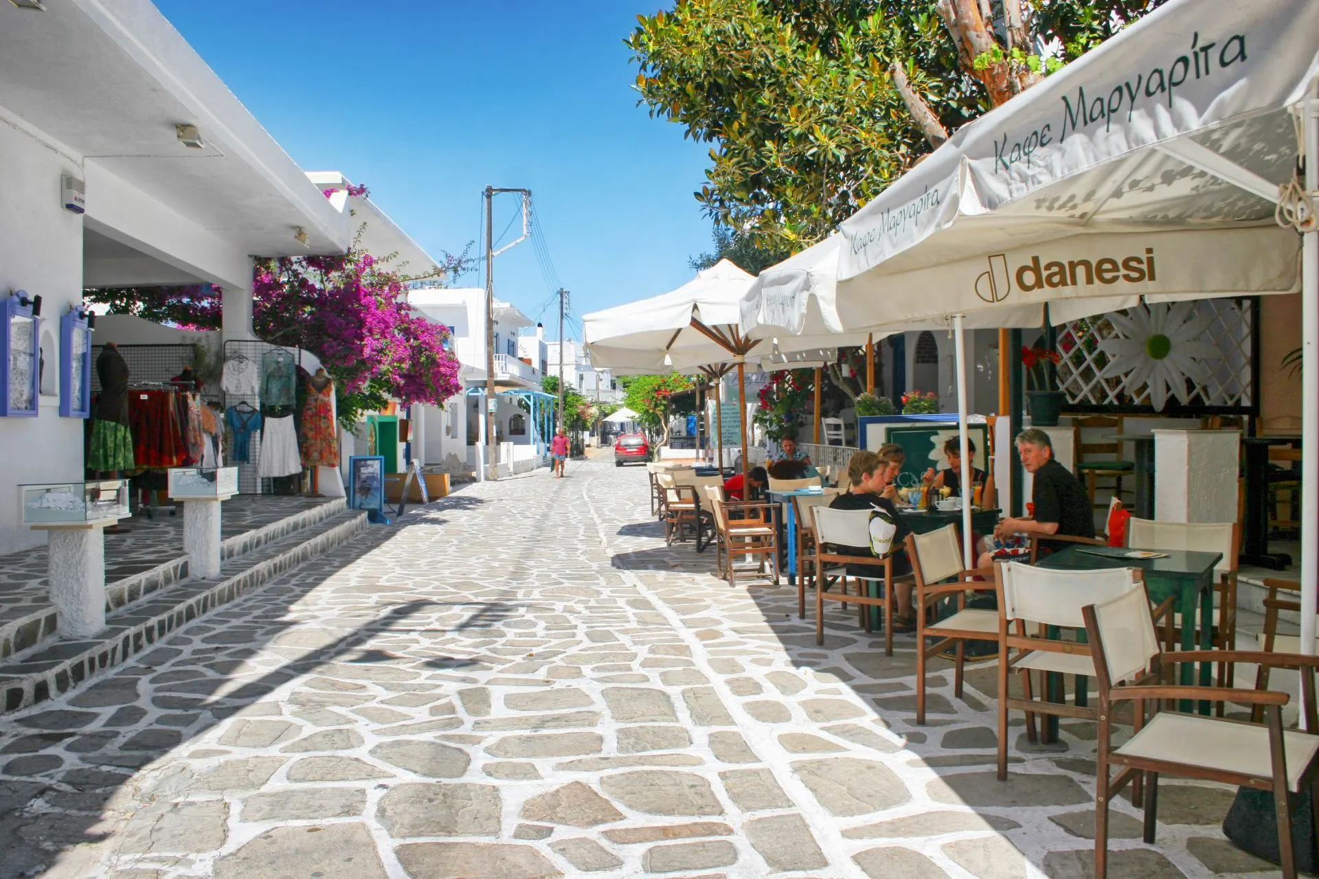 A vibrant street in Antiparos is lined with white-washed buildings and cafes, with purple bougainvillea flowers creating a beautiful archway.