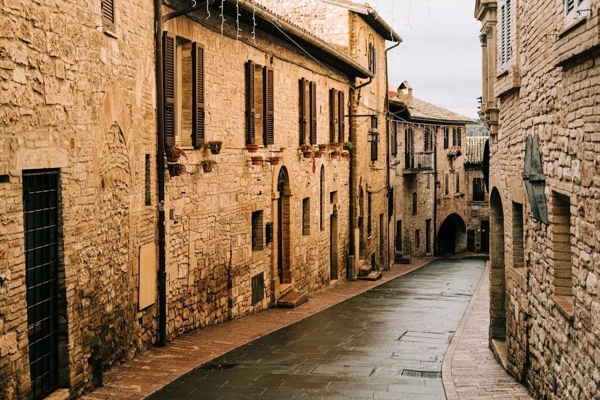 A narrow, cobblestone street winds between the historic stone buildings of Assisi, creating a charming and atmospheric scene.