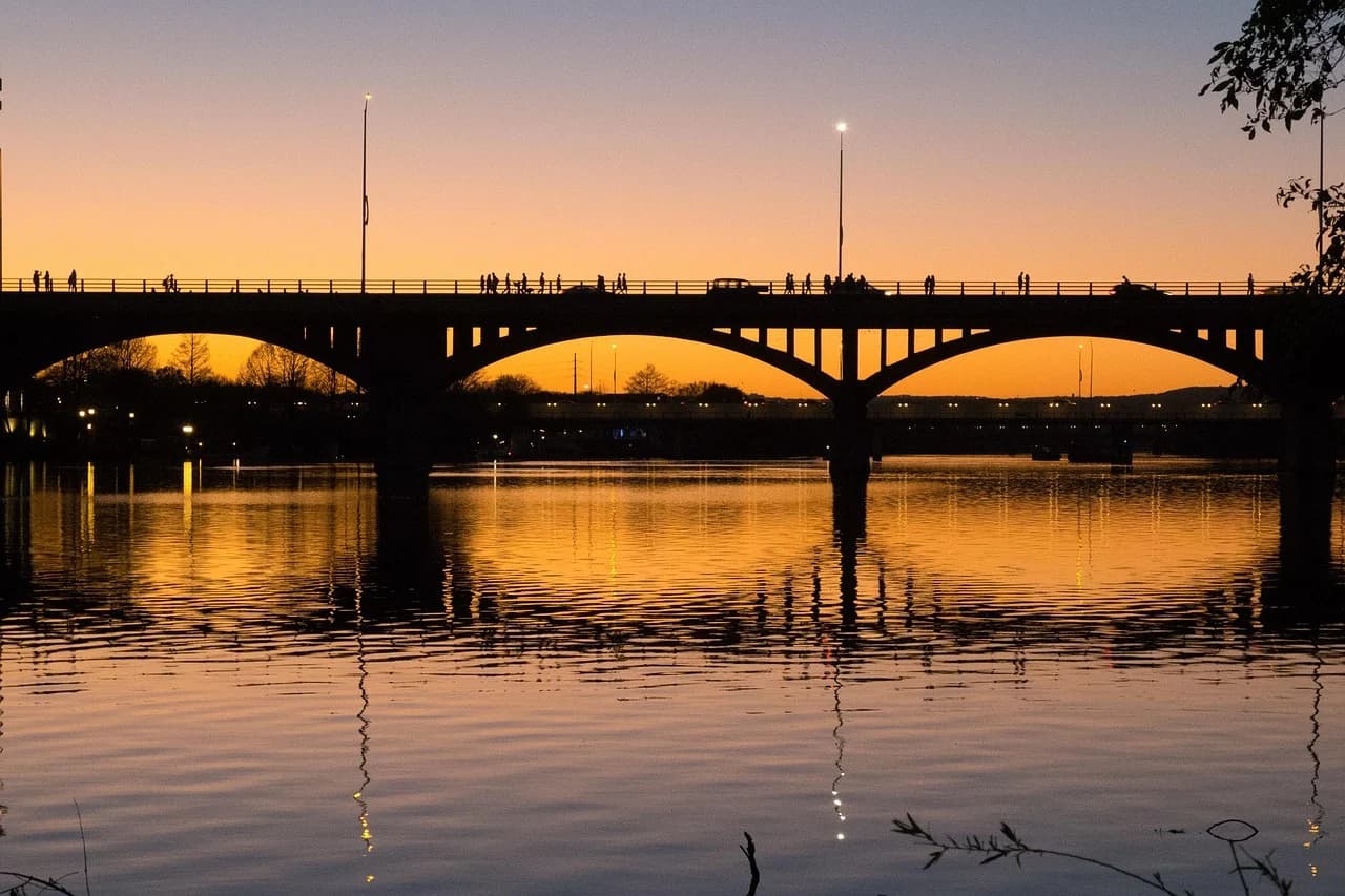 People walk and run along the Congress Avenue Bridge, which is silhouetted against a golden sunset reflected on the water.