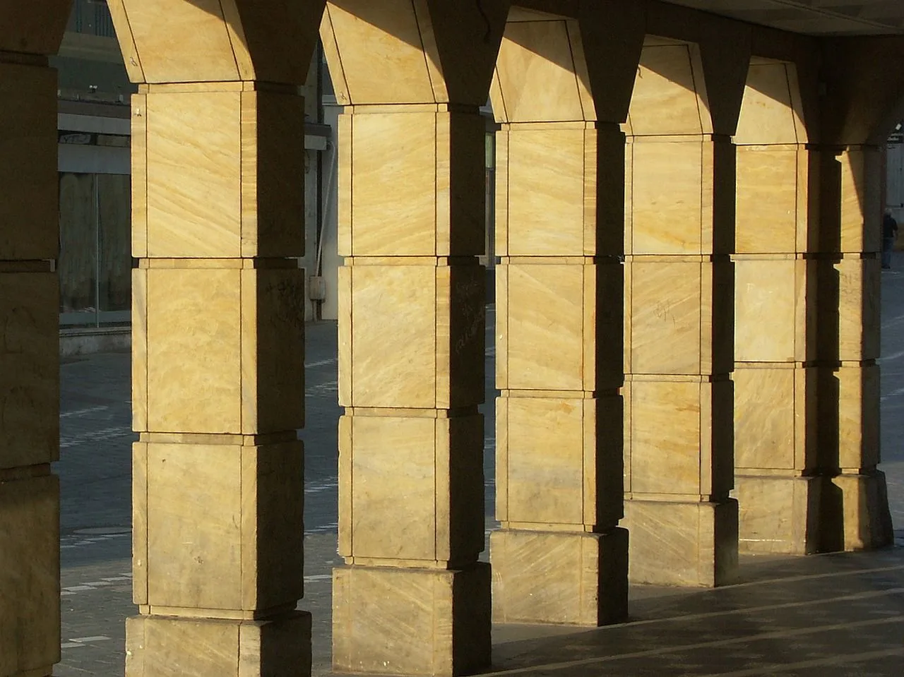 A series of large, square-shaped columns stand in a line, casting long shadows on a stone walkway.